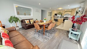 A modern living room with a brown leather sofa, a dining table with chairs, a kitchen area, and a potted plant.