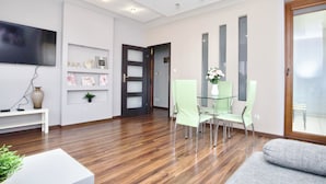 A modern living room with a wooden floor, a glass dining table with green chairs, a grey sofa, and a flat-screen TV mounted on the wall.