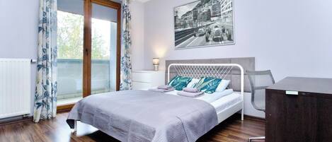 Un dormitorio con cama, escritorio, ventana con cortinas, radiador y un cuadro en la pared.