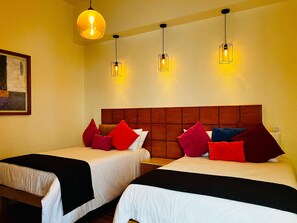 Ein Hotelzimmer mit zwei Betten, jeweils mit einem hölzernen Kopfteil, weißer Bettwäsche und mehreren roten und blauen Kissen. Drei Pendelleuchten hängen von der Decke.