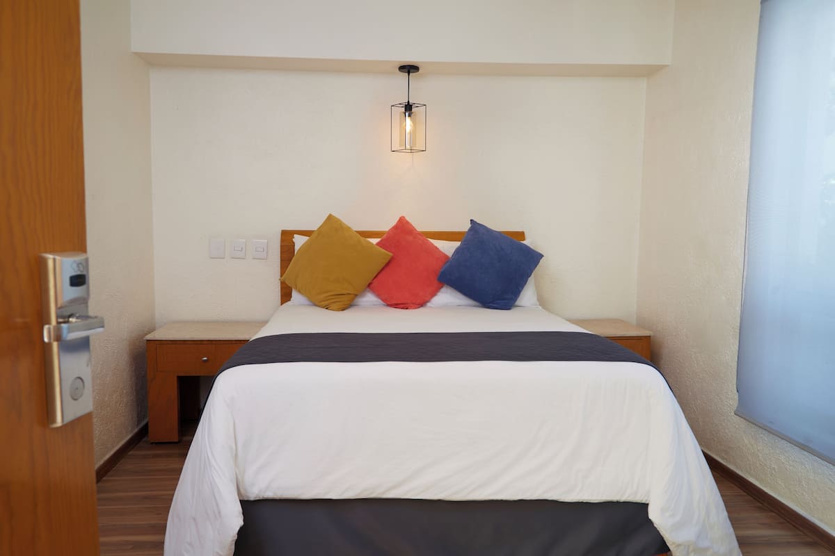Hotellihuone, jossa on sänky, yöpöytä, seinään kiinnitetty valaisin ja ikkuna.