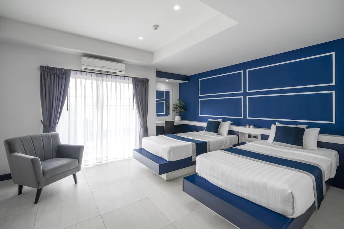 Quarto de hotel moderno com duas camas, uma poltrona cinza e uma janela grande com cortinas.