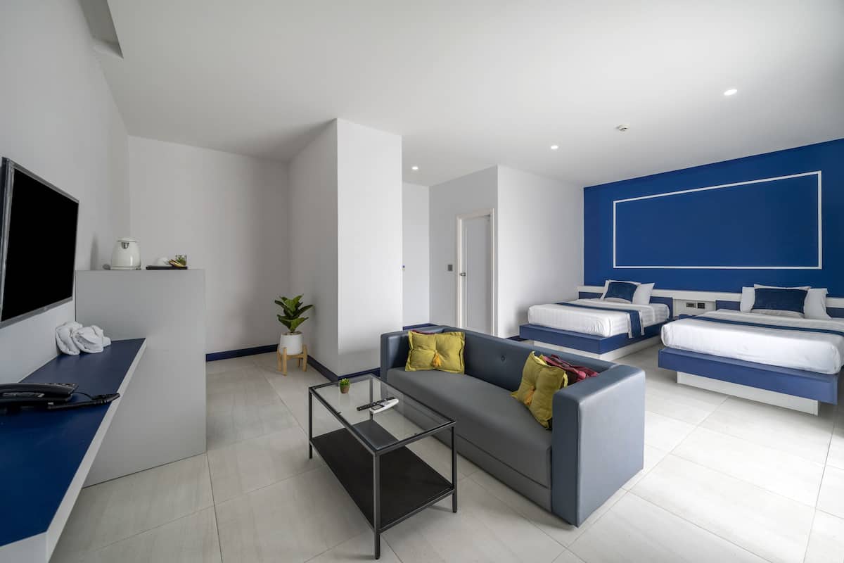 Quarto de hotel moderno com sofá, mesa de centro de vidro, televisão e cama com cabeceira azul.