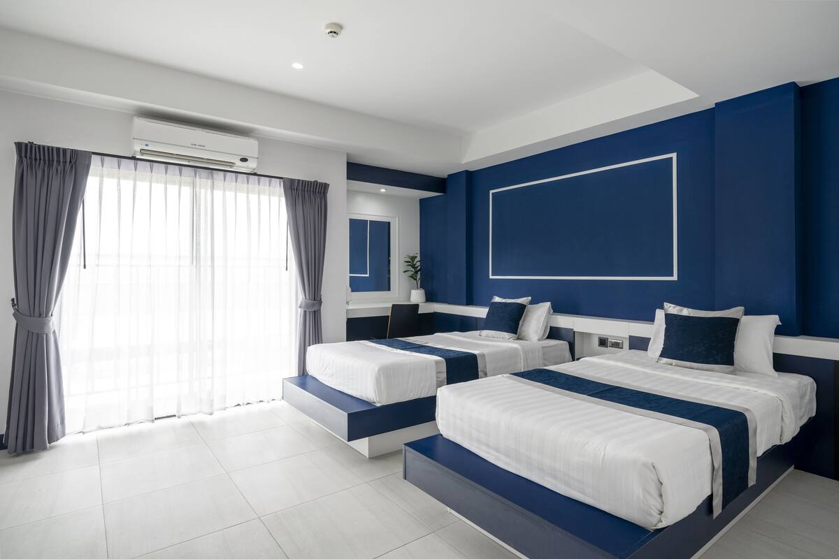 Quarto de hotel moderno com duas camas, uma janela grande com cortinas, ar condicionado e uma parede azul de destaque.