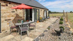 A stone building with a patio area featuring a table and chairs, a red umbrella, and a stone pathway leading to the building. - The Cottage (Chesterfield)