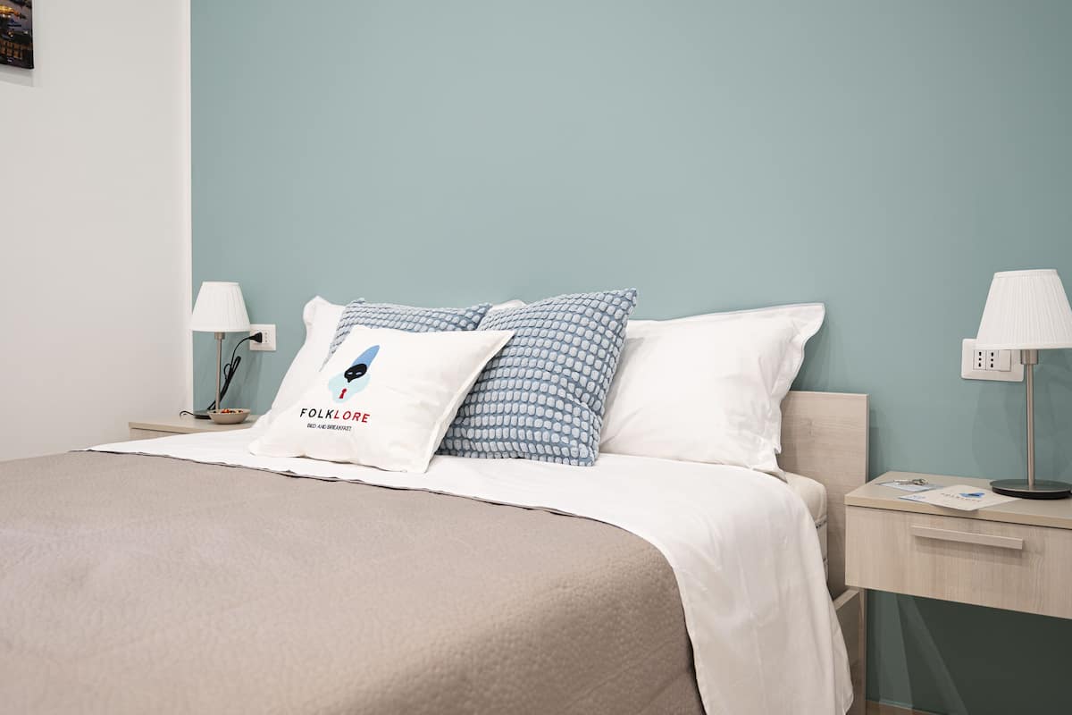 A neatly made bed with a beige comforter and white pillows, a bedside table with a lamp, and a wall-mounted power outlet.