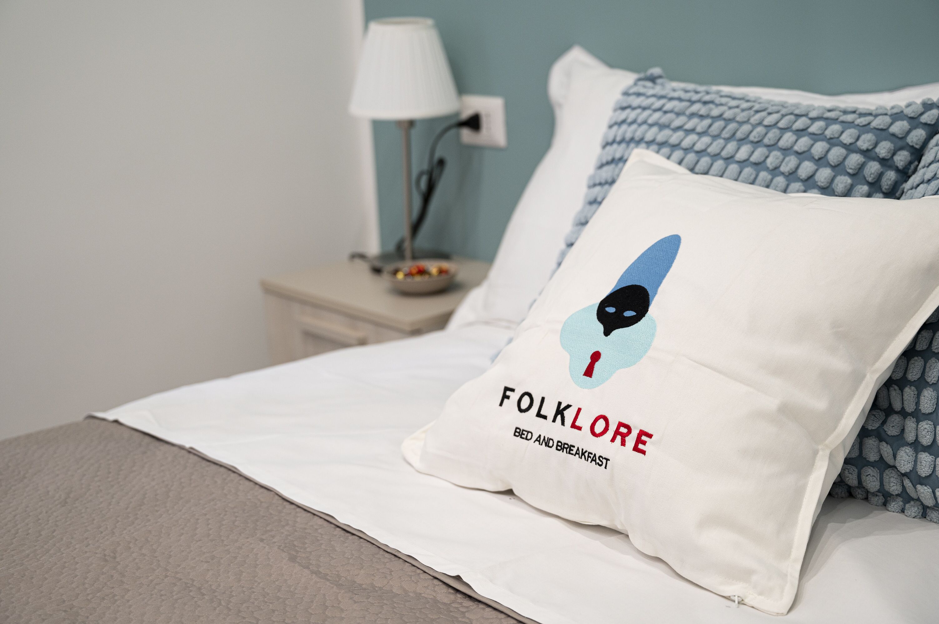 A bed with a white pillow featuring a logo and text, a blue patterned pillow, a bedside table with a lamp, and a bowl of fruit.