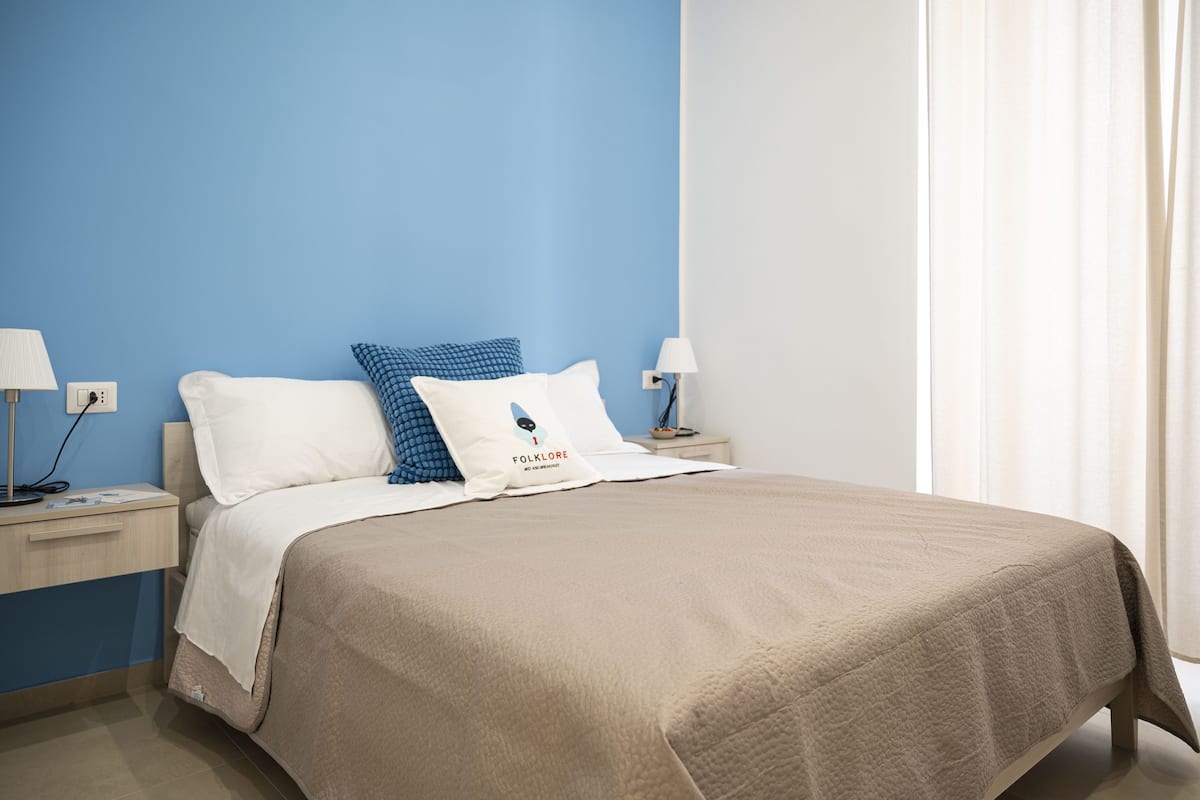 A neatly made bed with a beige cover, two pillows, one with a blue pattern, and a white pillow with a logo. There are two bedside tables with lamps, one on each side of the bed.
