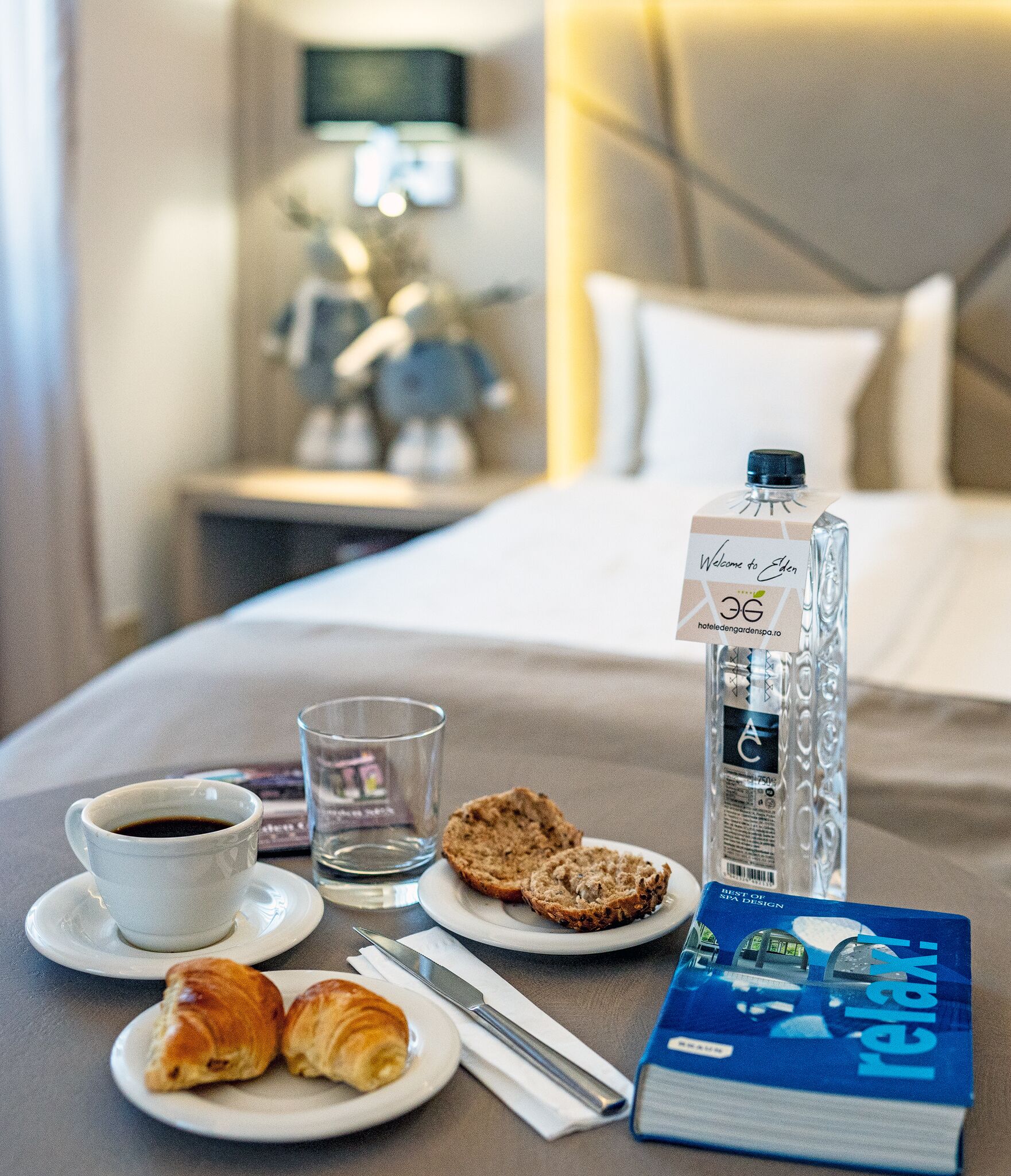 Ein liebevoll gedeckter Frühstückstisch auf einem Hotelbett mit einer Wasserflasche, einer Tasse Kaffee, Croissants und einem Buch.
