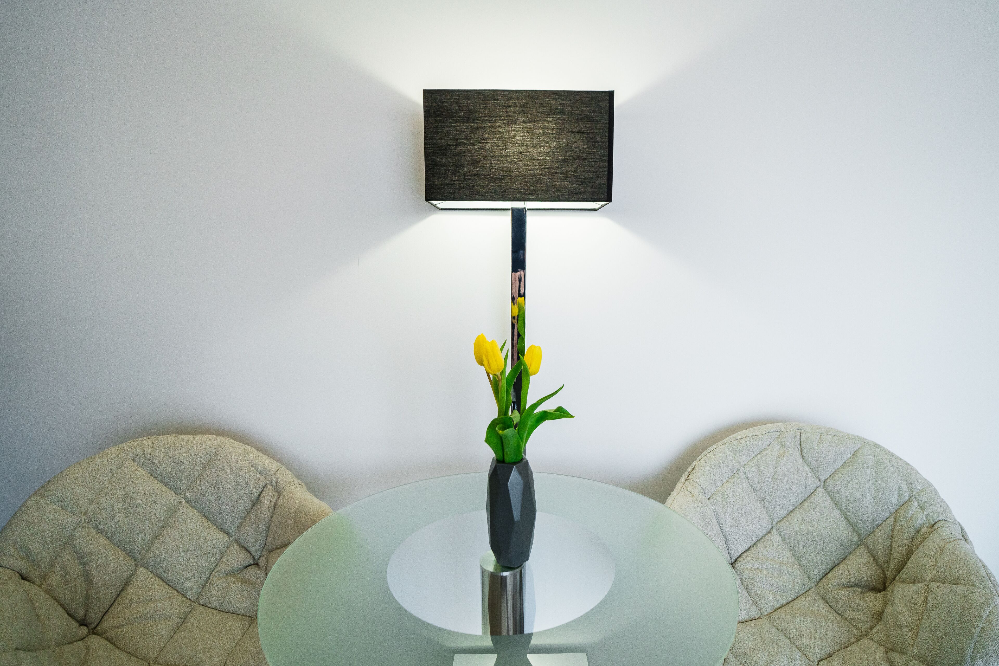 A modern dining area with a round glass table, a vase with yellow flowers, and two cushioned chairs.