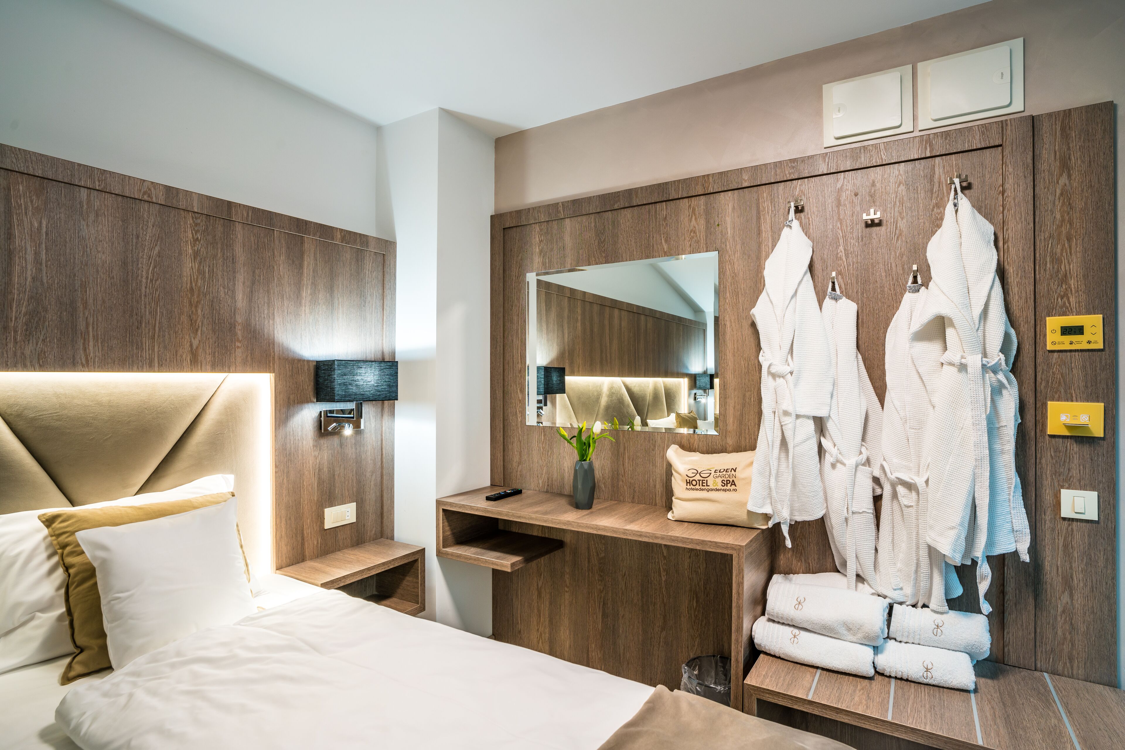 Ein modernes Hotelzimmer mit einem Bett, einem Schminktisch mit Spiegel, einem Bademantelständer und einem kleinen Schreibtisch mit Blumen in einer Vase.