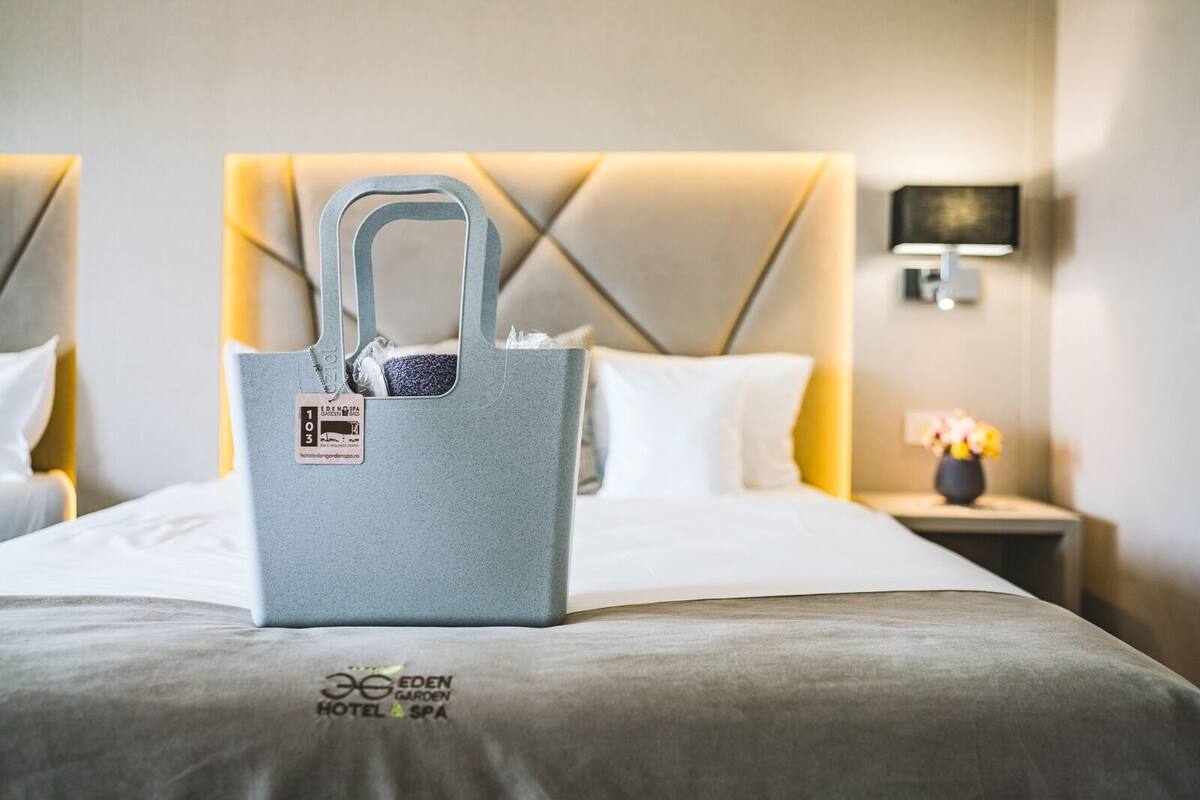 Habitación de hotel con cama, mesita de noche con lámpara y una bolsa gris sobre la cama.