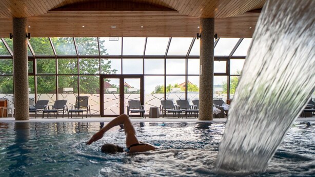 Una piscina con caída de agua, sillas de descanso y ventanas amplias.