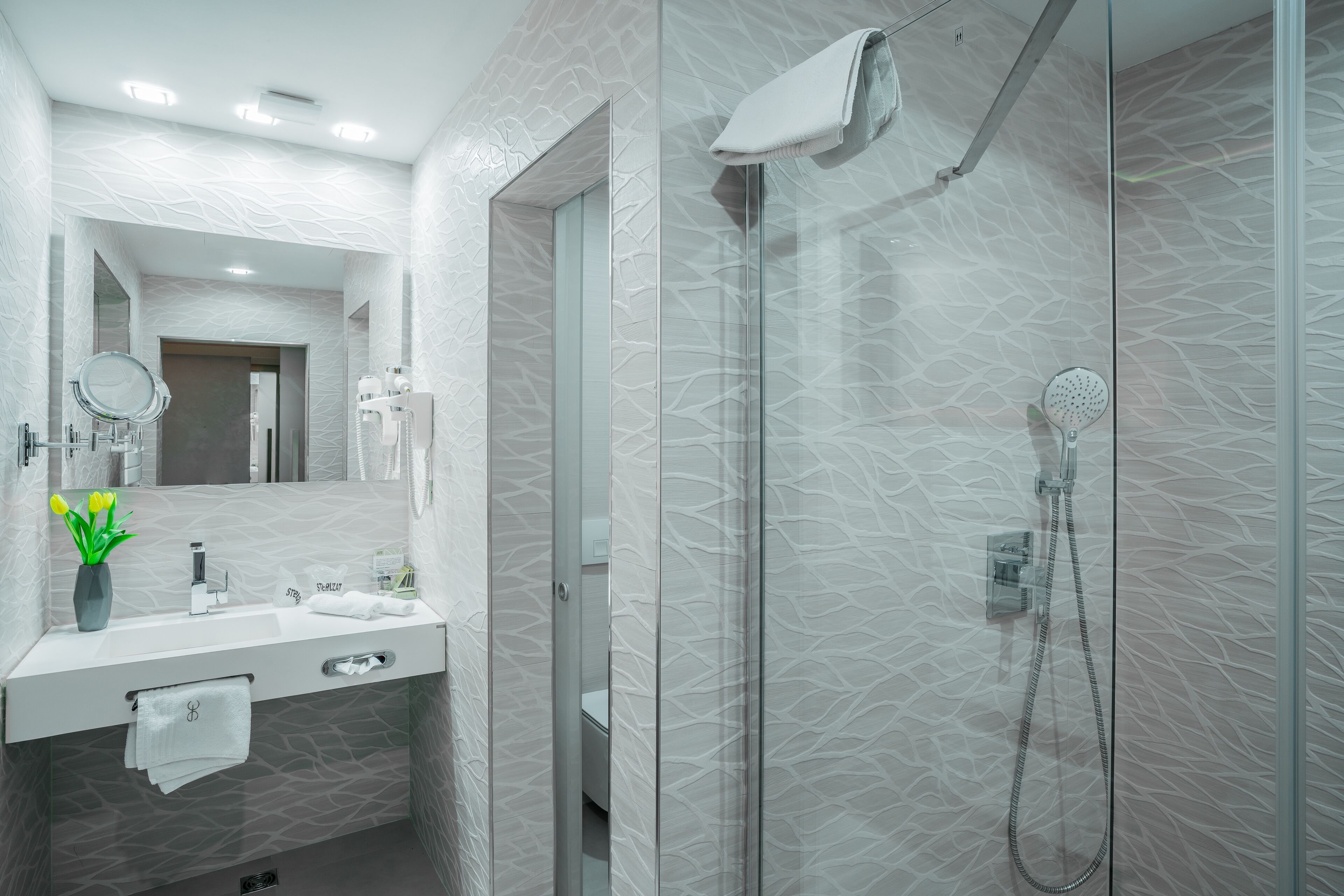 Un baño moderno con ducha de vidrio, tocador con espejo y un jarrón con flores amarillas.