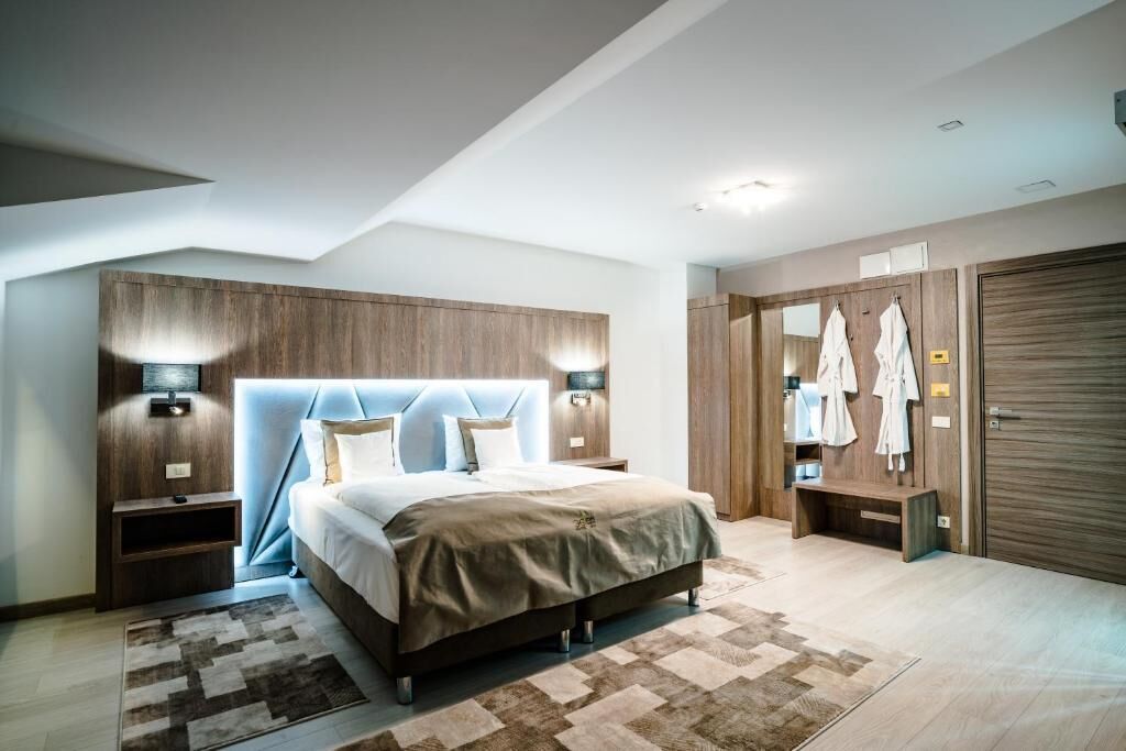 Un dormitorio moderno con una cama grande, cabecera de madera, lámparas de noche, un banco y un armario con ropa colgada.