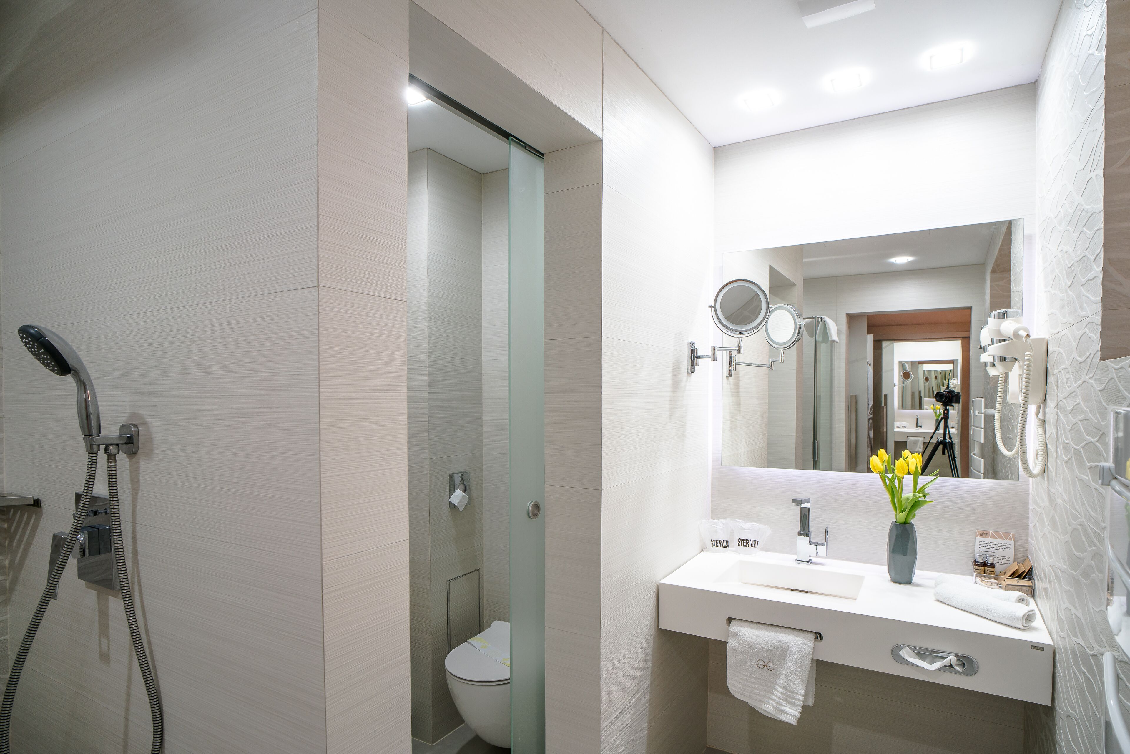 Un baño moderno con ducha de vidrio, lavamanos blanco, espejo y un florero con flores.
