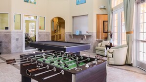 A pool table and foosball table in a well-lit room with a large window, a sofa, and a decorative wall piece. - Harmony Haven at Watersong (Davenport)