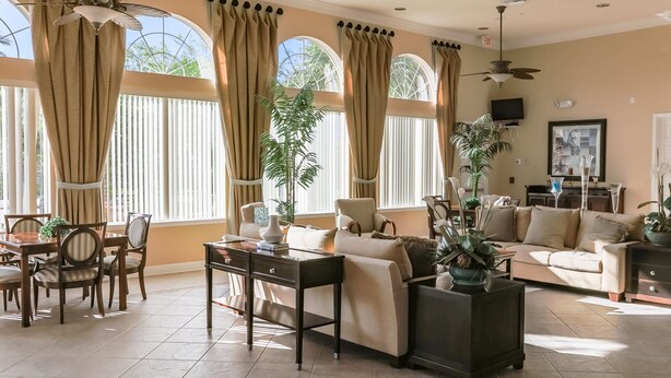 A spacious living room with a dining area, large windows with curtains, a ceiling fan, and a television. - Harmony Haven at Watersong (Davenport)