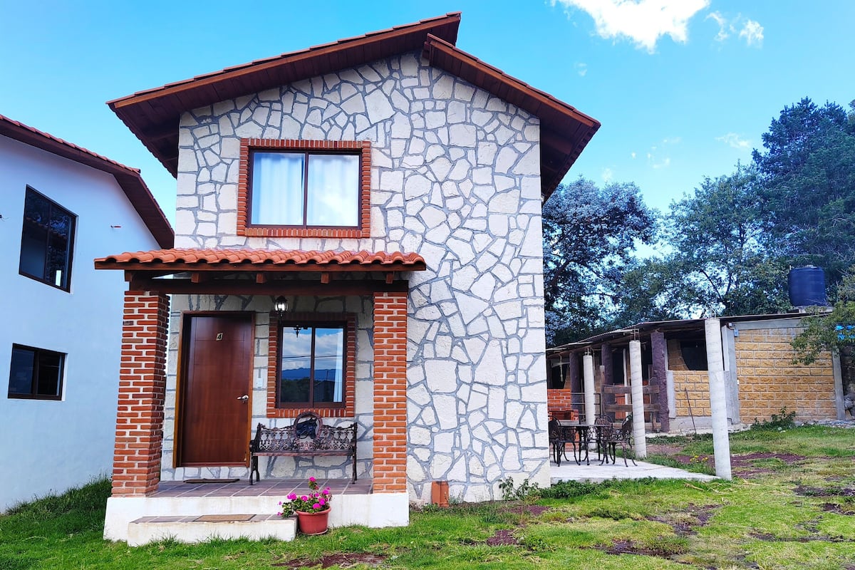Una casa de dos pisos con fachada de piedra, tejado de tejas y un porche delantero con un banco y plantas en macetas.