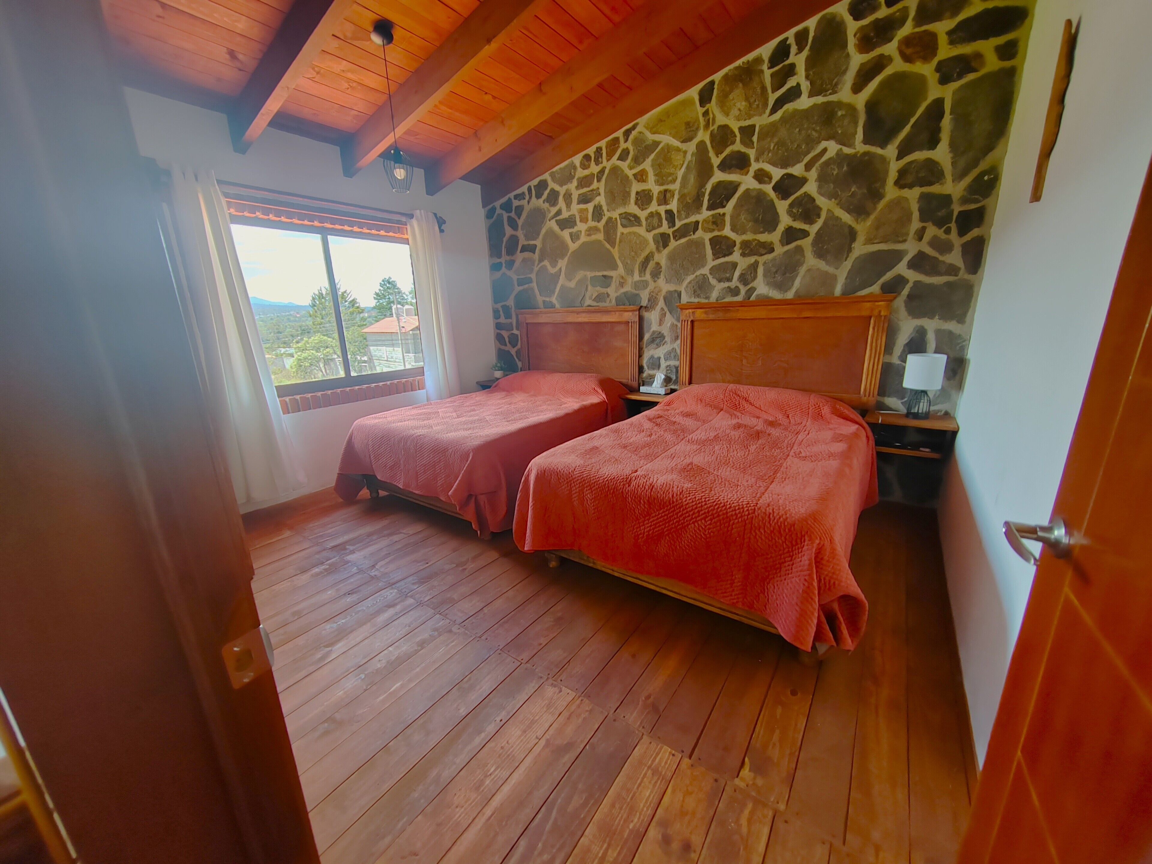 A bedroom with two beds, wooden flooring, a stone wall, and a window with a view.