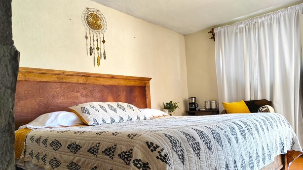Ein ordentlich bezogenes Bett mit gemusterten Bettwäsche, ein Holzkopfschild, ein an der Wand befestigter Traumfänger, eine Kaffeemaschine und ein Topf mit einer Pflanze auf einem Nachttisch.