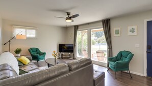 Een woonkamer met een grijze bank, twee groene fauteuils, een flatscreen televisie, een plafondventilator en een raam met jaloezieën.