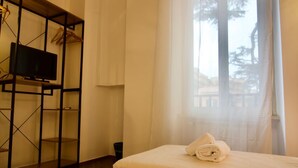 A hotel room with two beds, a TV on a shelf, and a window with sheer curtains.