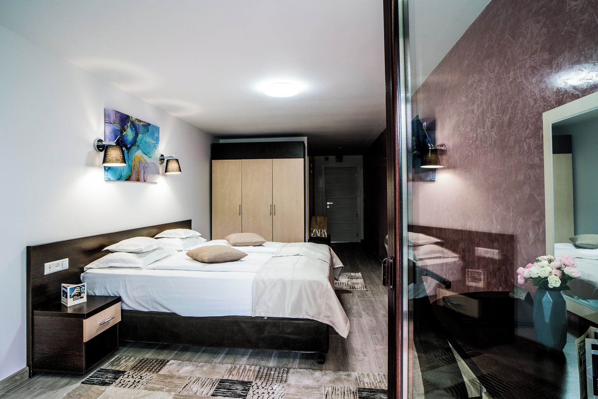 Un dormitori modern amb un llit, mobles de nit, un armari i un bany visible a través d'una porta de vidre.