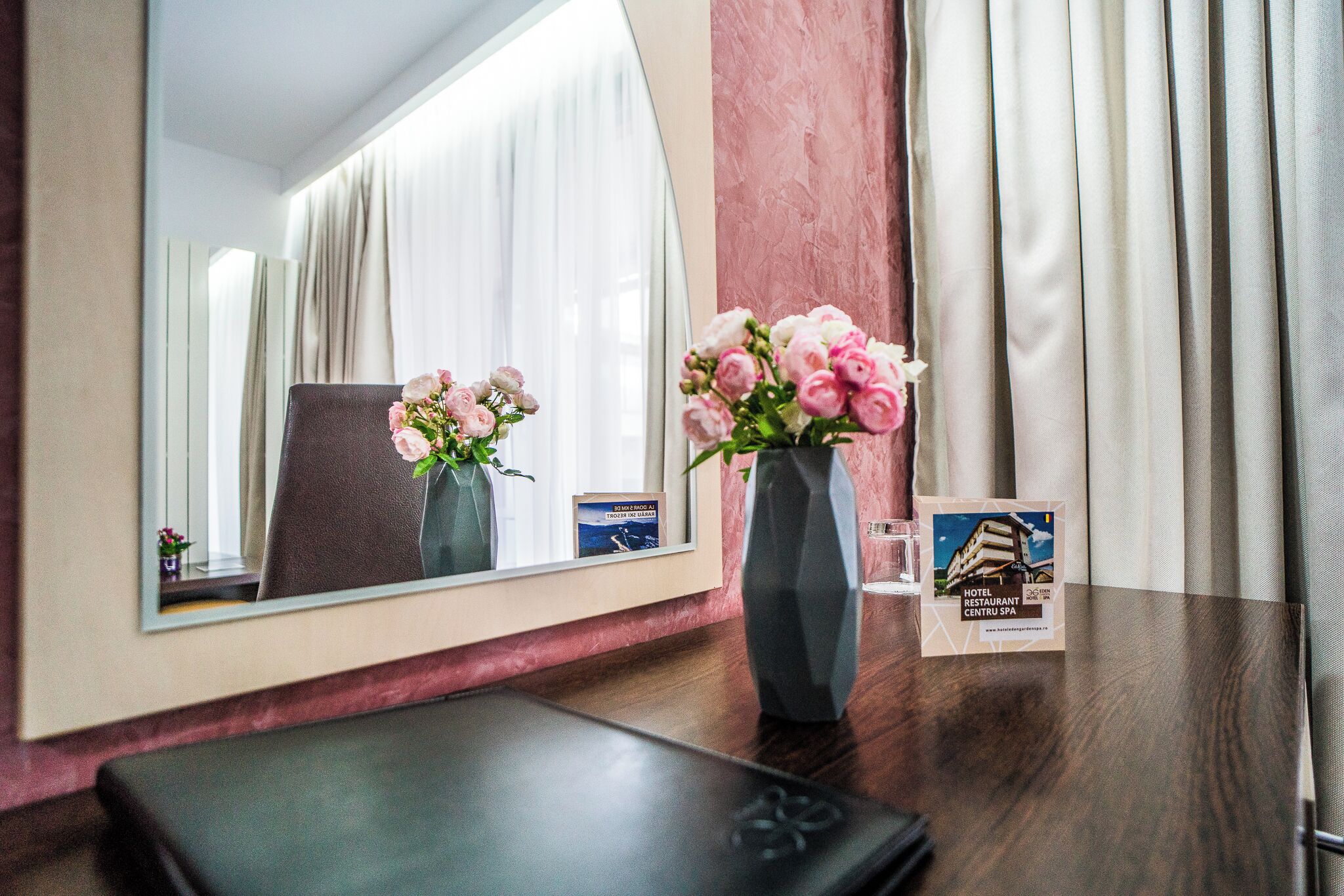 A hotel room with a wooden desk, a vase with pink flowers, a framed mirror, and a brochure on the table.