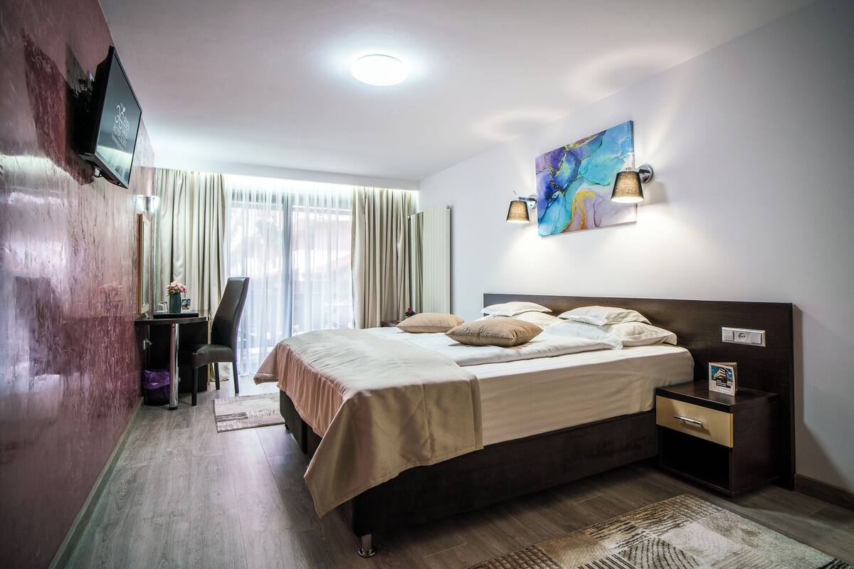A hotel room with a bed, bedside table, lamp, artwork, and a TV mounted on the wall.