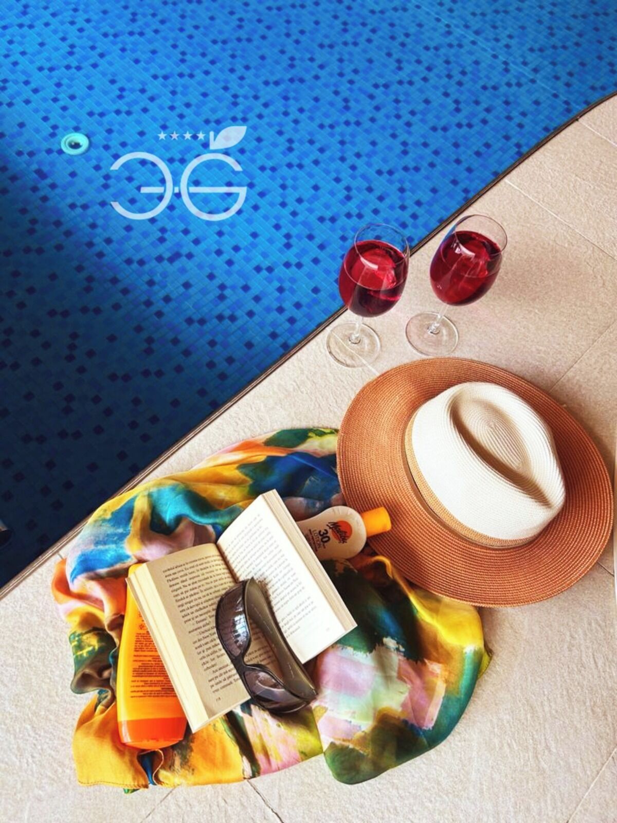 Une scène de bord de piscine avec un sol carrelé bleu, un chapeau de paille, un livre, des lunettes de soleil et deux verres de vin rouge.
