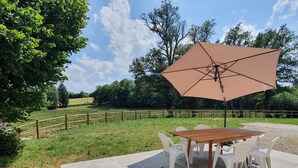 Een terras buiten met een houten tafel en witte plastic stoelen, beschaduwd door een grote parasol.