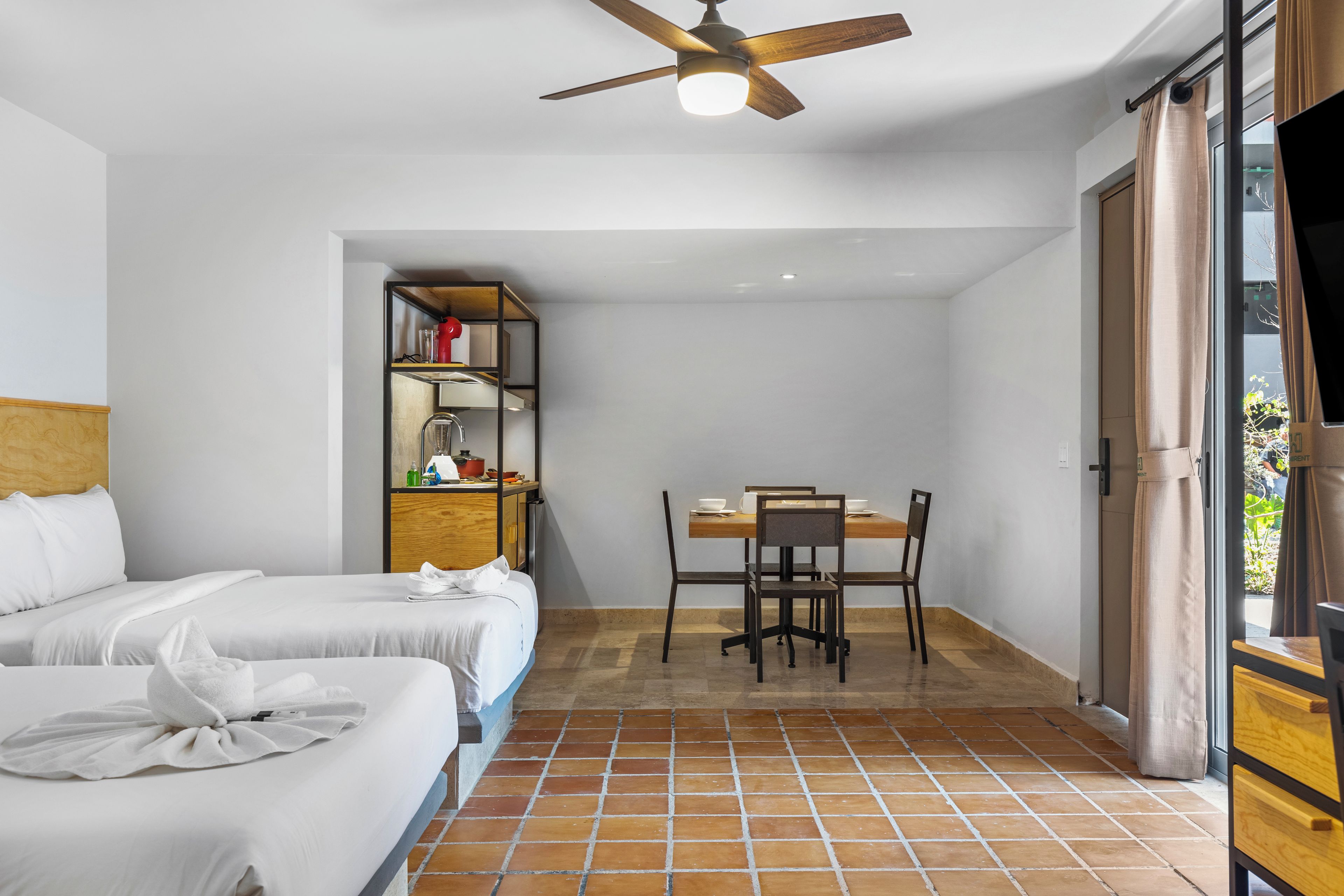 Quarto de hotel com duas camas, uma mesa de jantar com cadeiras, um ventilador de teto e vista para o exterior pela janela.