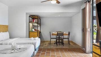 Quarto de hotel com duas camas, uma mesa de jantar com cadeiras, um ventilador de teto e vista para o exterior pela janela.