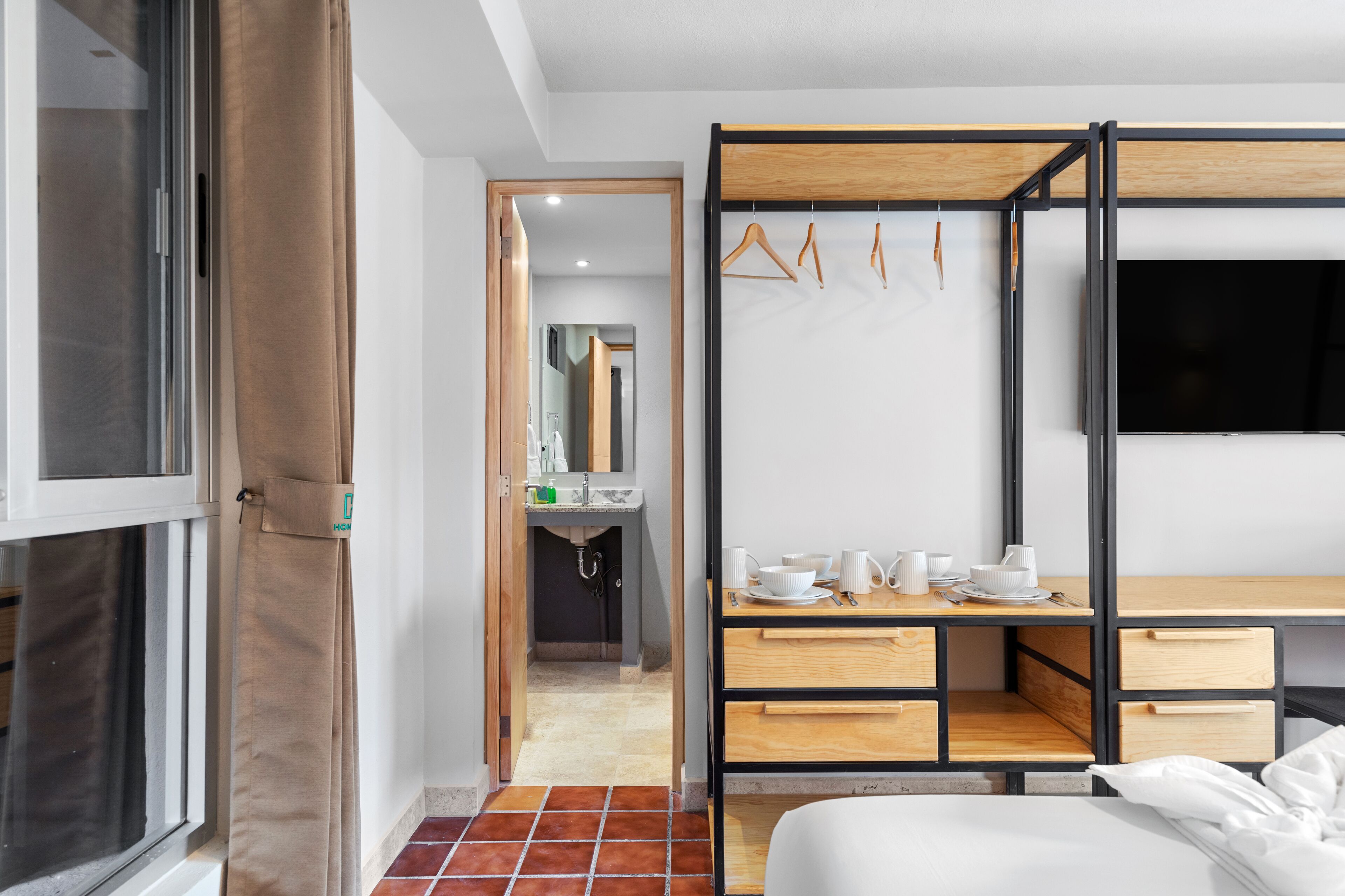 A compact hotel room with a bed, a wooden desk, a mirror, and a wardrobe with a TV.