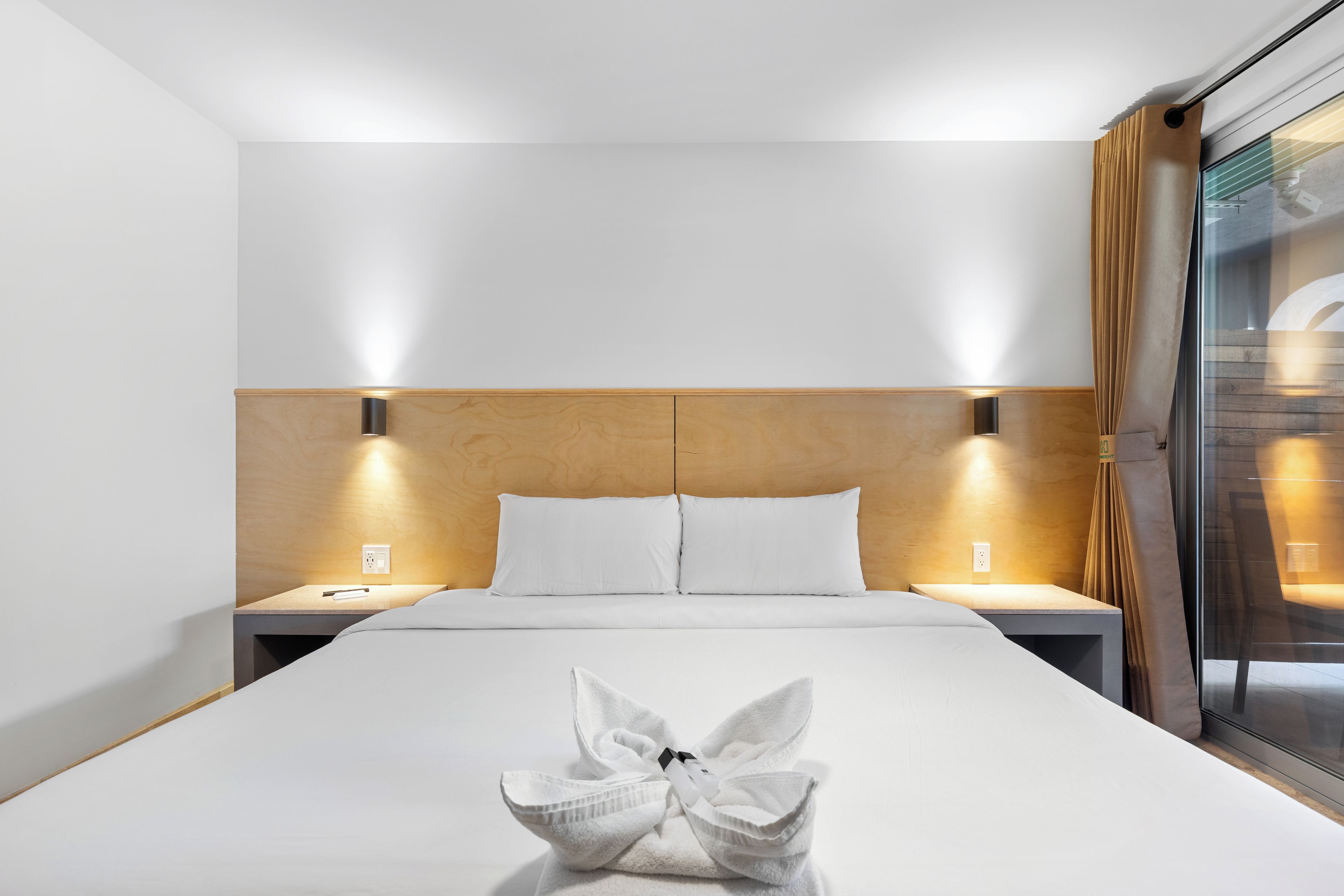 Une chambre d’hôtel avec un grand lit, deux tables de chevet, une tête de lit en bois et une fenêtre avec des rideaux.