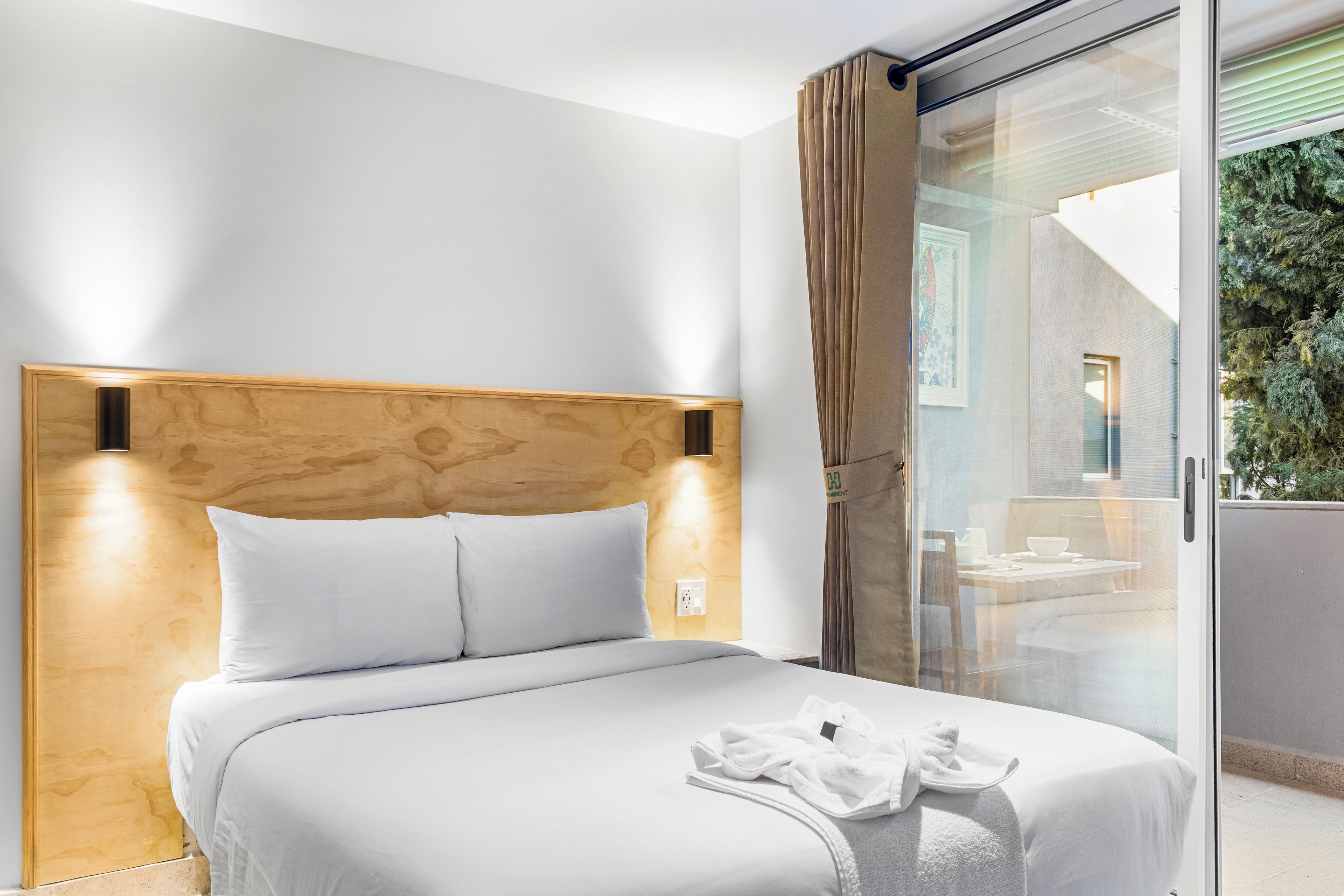 A hotel room with a wooden headboard, a bed with white bedding, and a glass door leading to a balcony with a view of greenery.