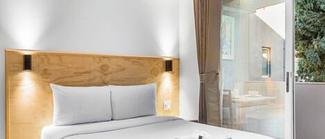 A hotel room with a wooden headboard, a bed with white bedding, and a glass door leading to a balcony with a view of greenery.