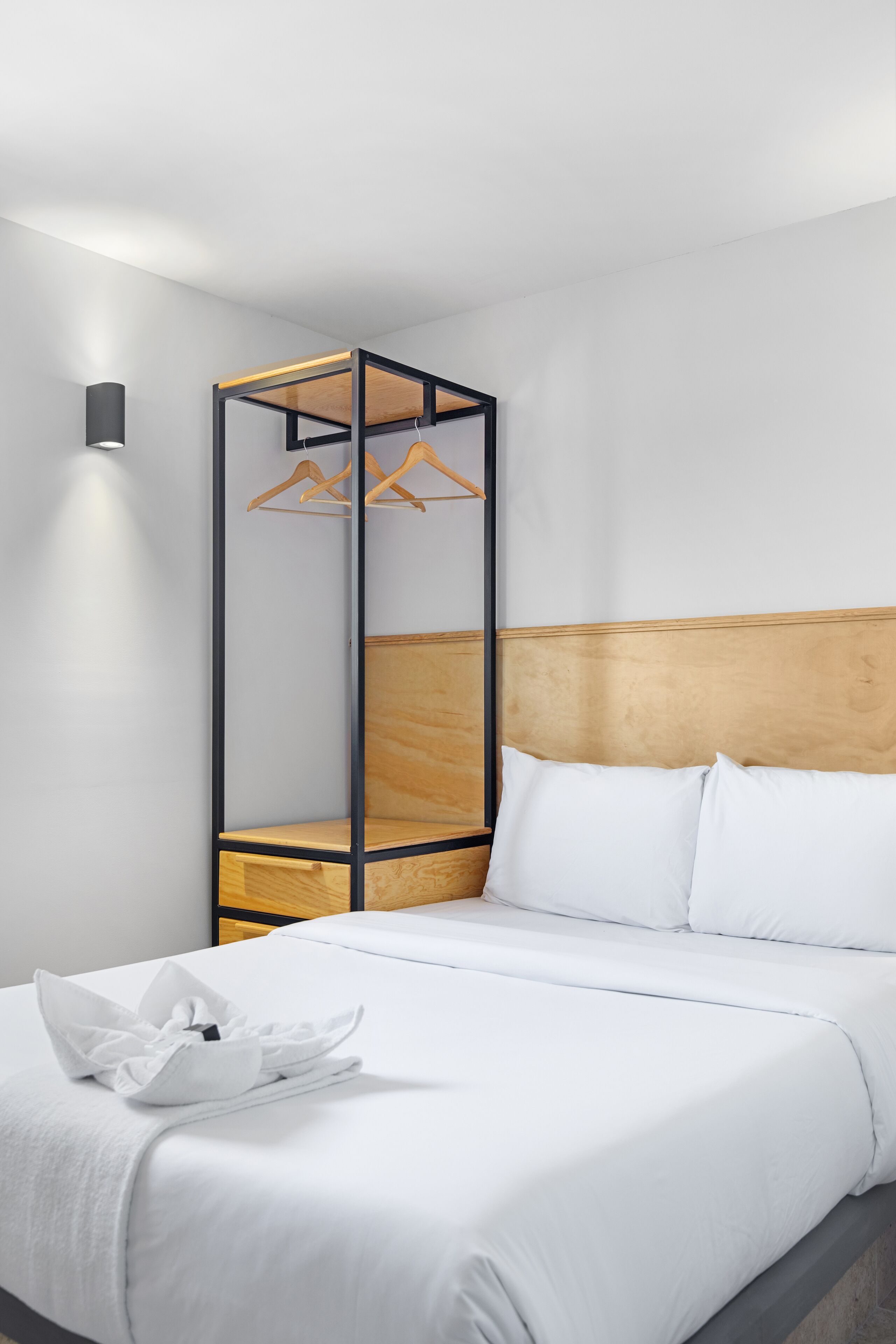 A modern hotel room with a neatly made bed, a wooden headboard, and a tall, narrow wardrobe with wooden shelves and a hanging rack.