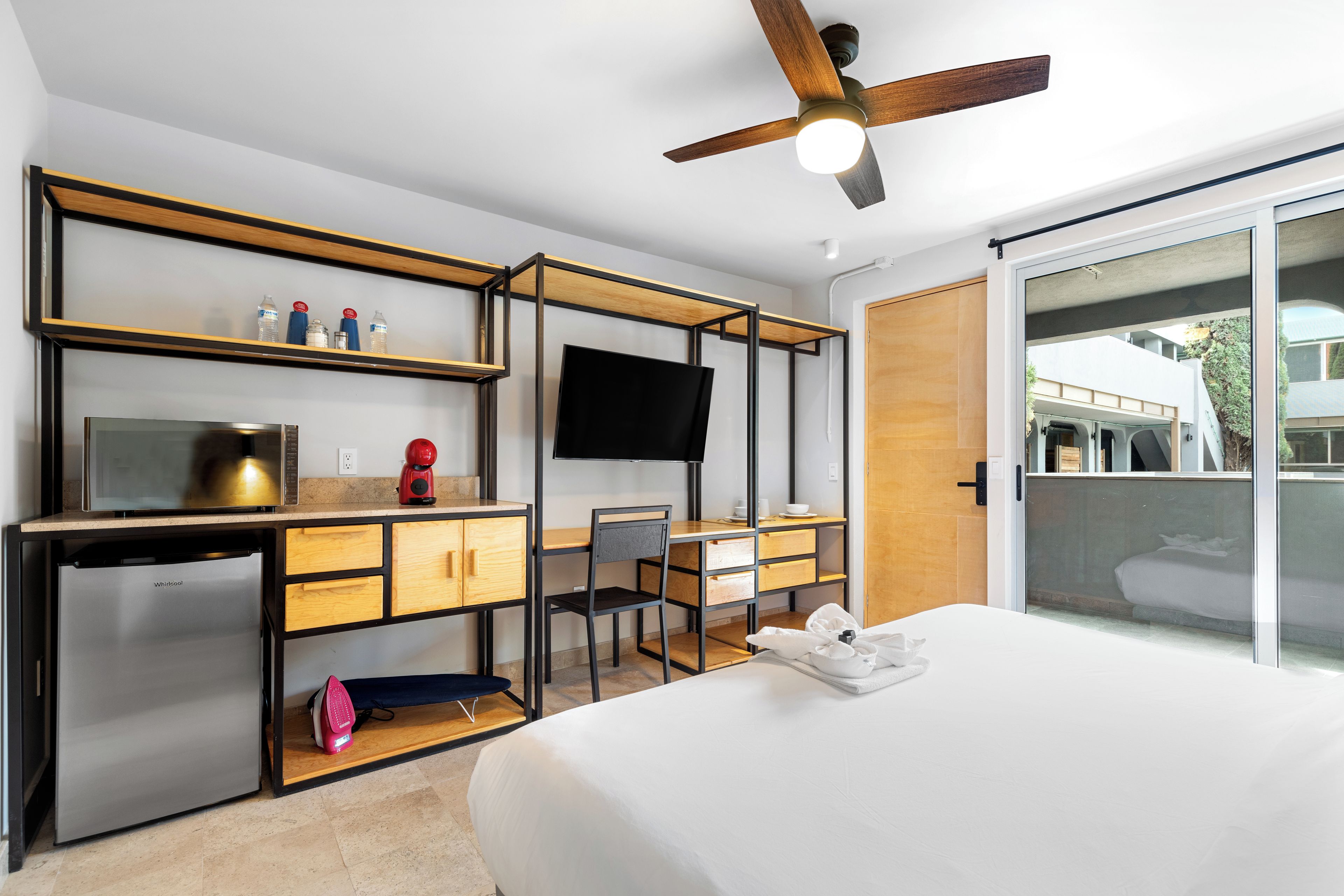 A modern hotel room with a bed, a desk with a chair, a television, a ceiling fan, and a sliding door to a balcony.