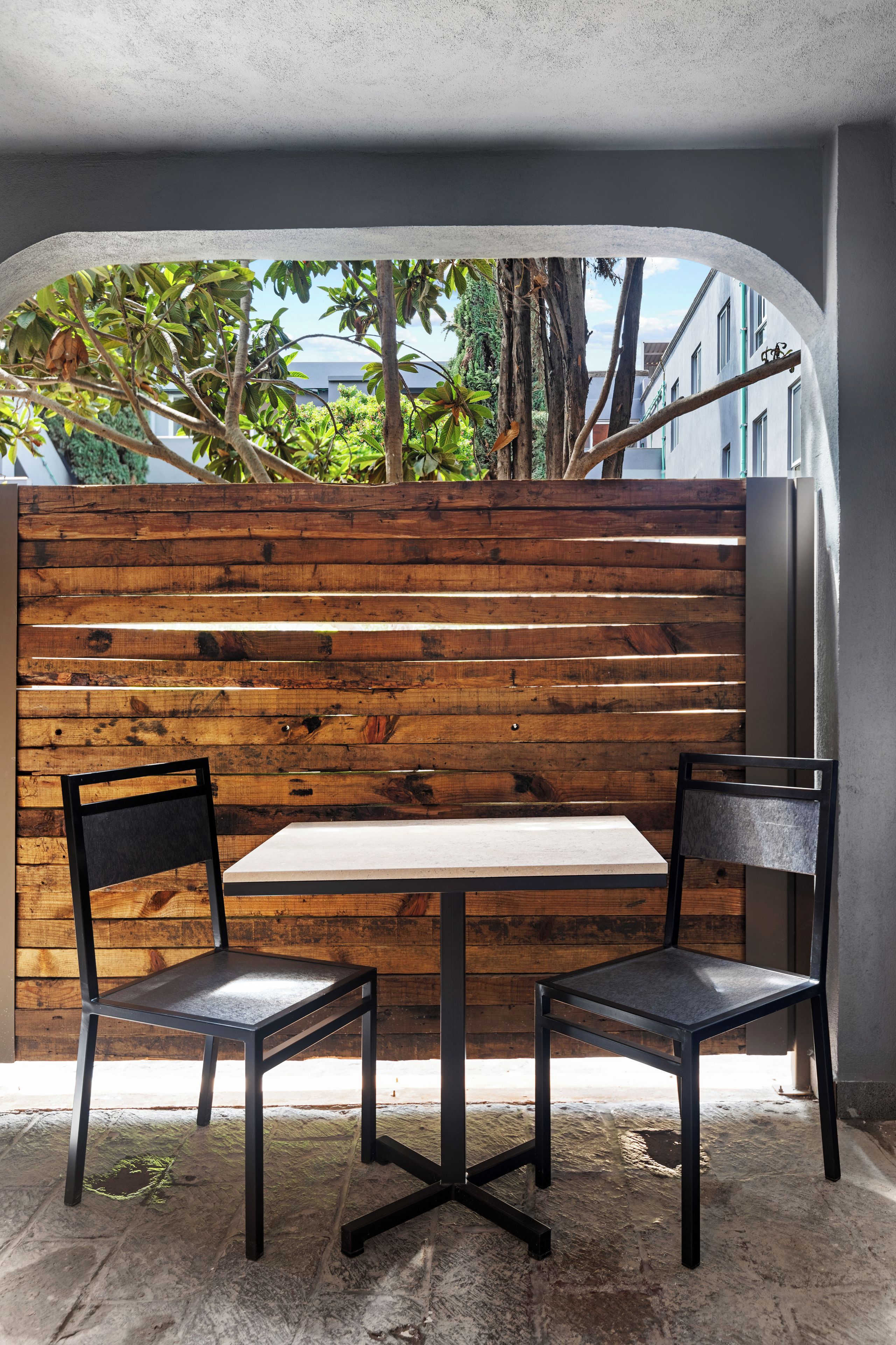 A table and two chairs in a covered outdoor area with a wooden partition and a view of greenery and buildings.
