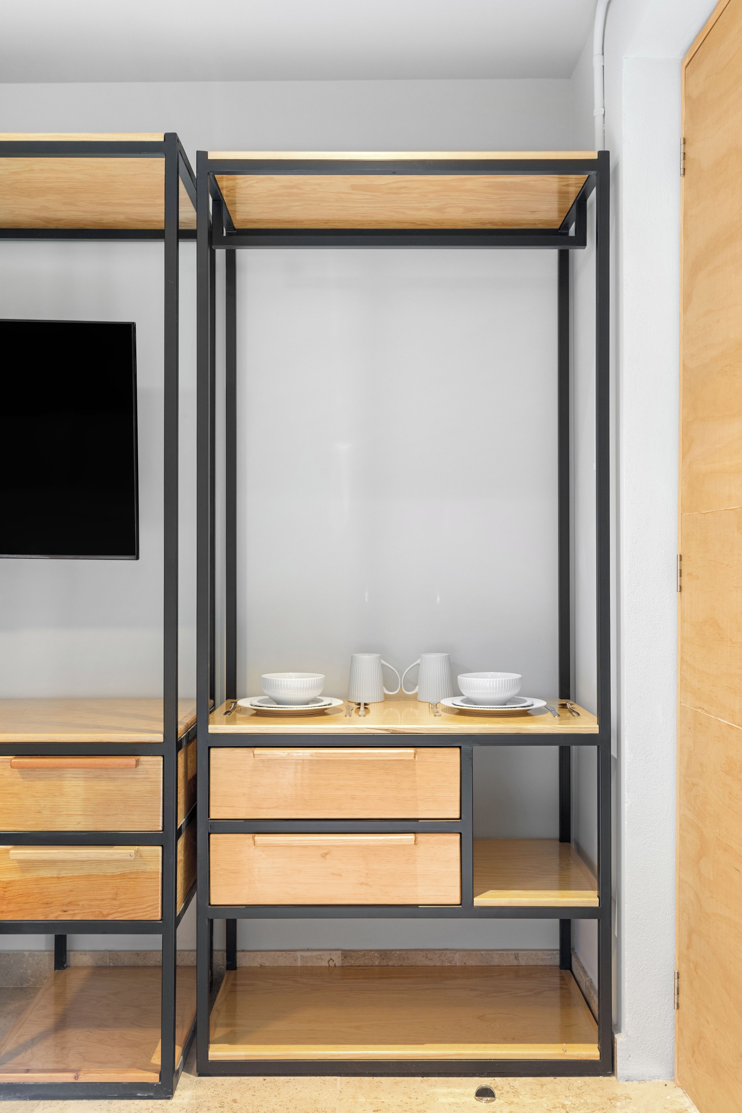 Una habitación compacta y moderna con escritorio integrado, superficie de madera y estructura metálica, televisor de pantalla plana en la pared y un pequeño rincón de estar con mesa y sillas.