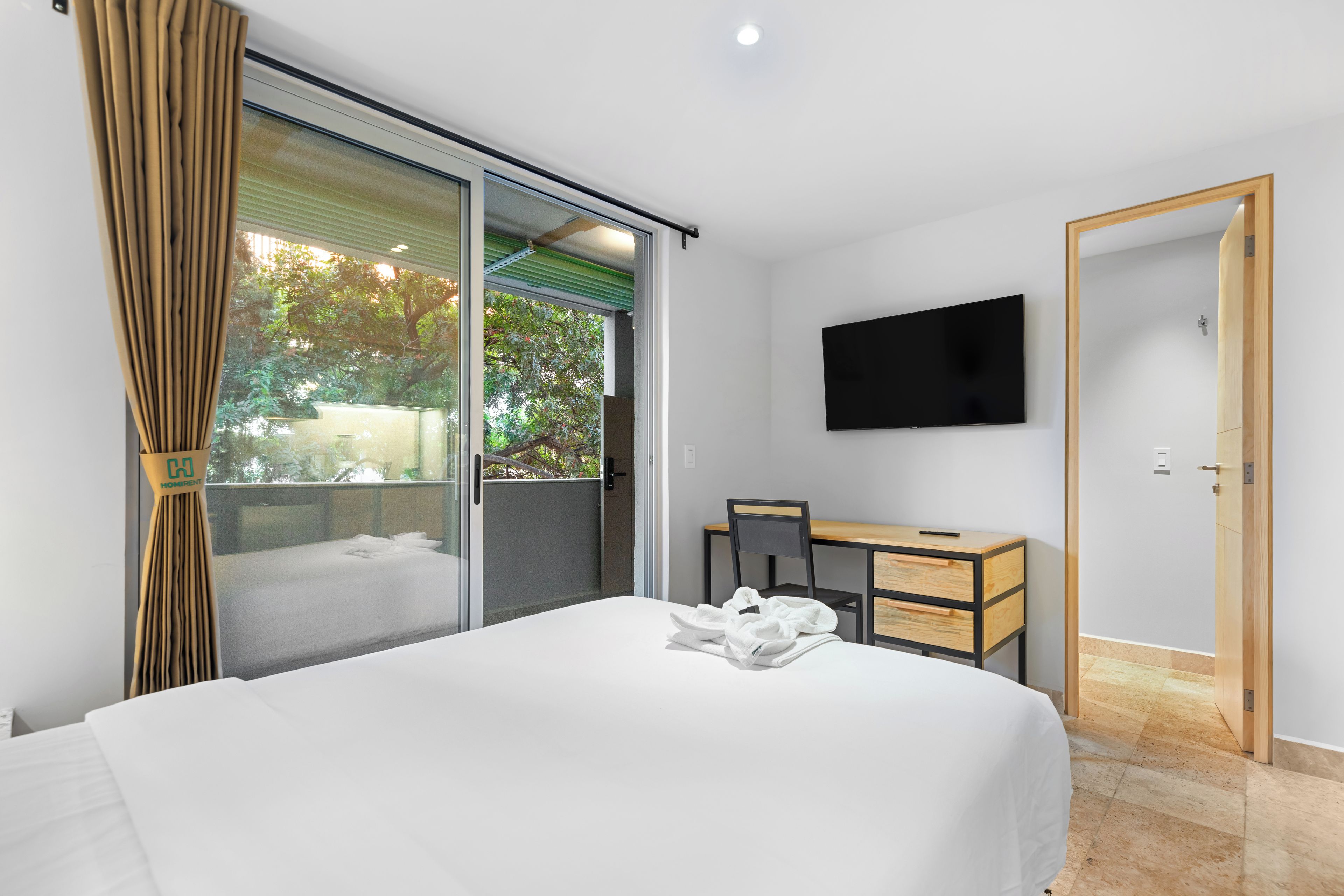 Quarto de hotel com cama, escrivaninha, cadeira, televisão e porta de correr para uma varanda.