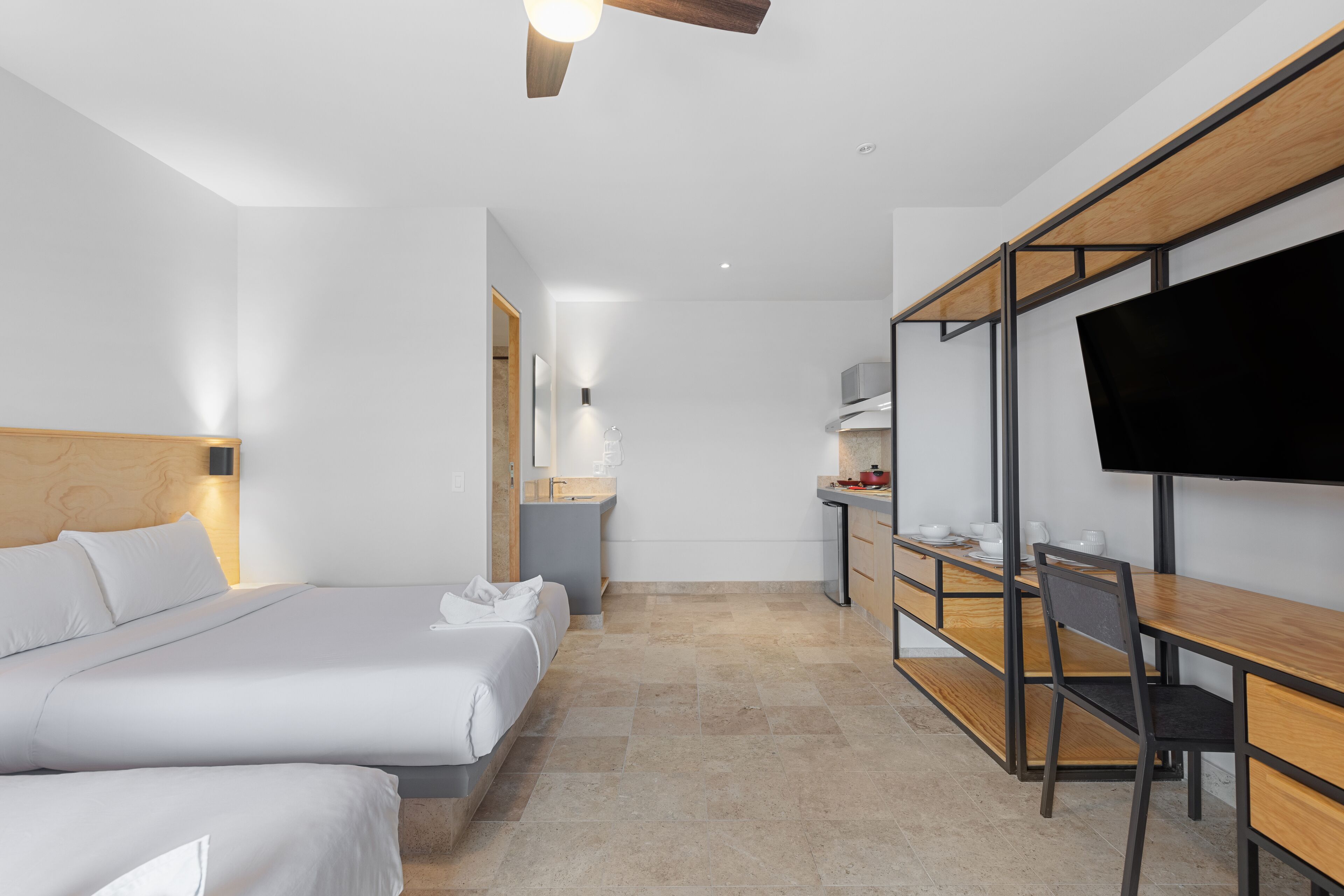 A modern hotel room with a large bed, a desk with a chair, a television, and a kitchenette.