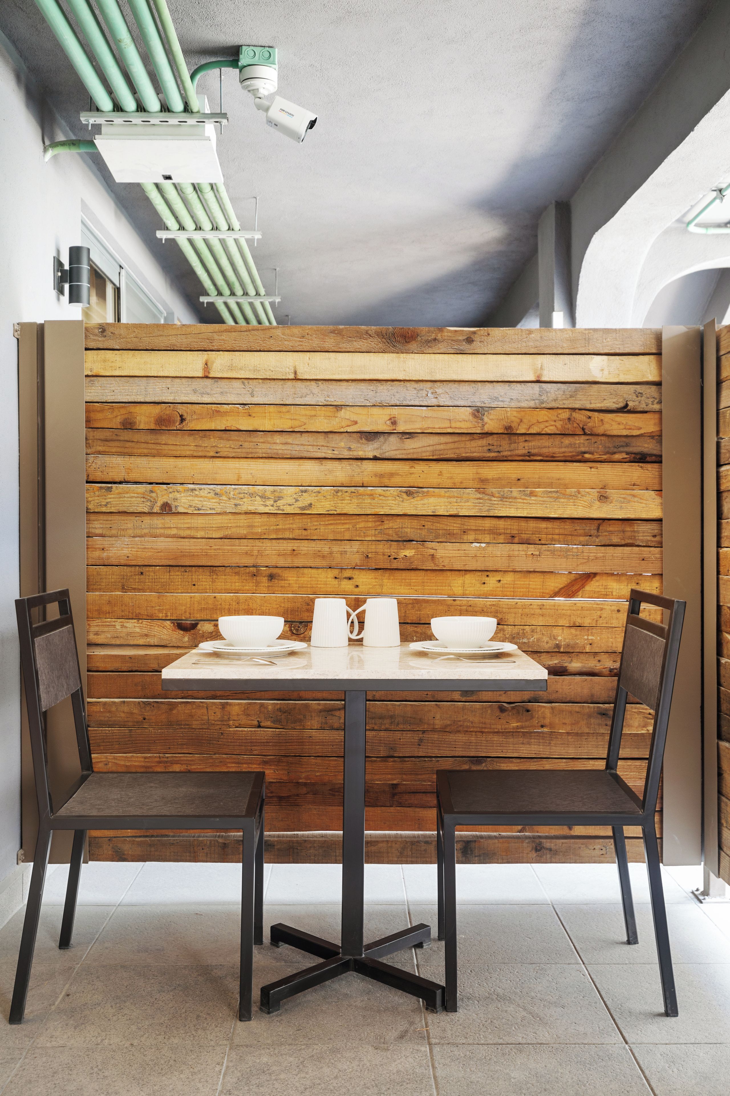 Ein Essbereich mit einer Holzwand, zwei Stühlen, einem Tisch mit weißem Geschirr und einer Deckenkamera.
