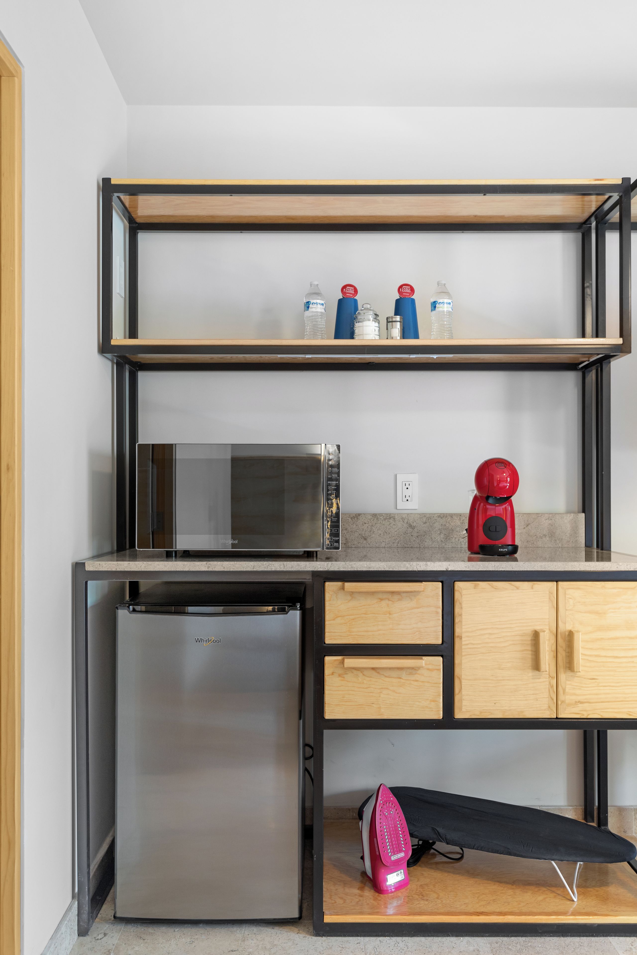 A compact kitchen with a stainless steel refrigerator, a microwave, a red coffee maker, and a shelf with water bottles.
