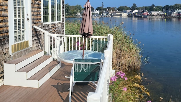 A wooden deck with a glass table and chairs, a hanging chair, and a view of a lake with buildings in the background.
