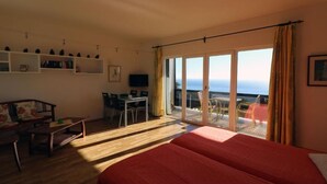 Ein Zimmer mit einem großen Bett, einem Schreibtisch, einem Sessel, einem Fernseher und einem Balkon mit Meerblick.