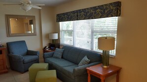 A living room with a sofa, two ottomans, a chair, a lamp, a phone, and a window with blinds. - Beachfront Condo with Private Balcony - Beachfront Resort- Pool - Kitchen  (St. Pete Beach)