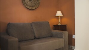A brown sofa with a cushion, a small table with a lamp, and a wall with a textured artwork. - R. Virgínia 48 (Búzios)
